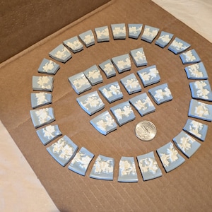 May include: A collection of light blue and white ceramic fragments arranged in a circular pattern with a smaller cluster in the center. Each piece features a raised white floral relief design. A silver-colored coin is placed near the center.