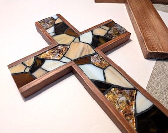May include: A wooden cross with a mosaic design. The mosaic features a mix of brown, tan, white, and gold-colored glass pieces. The cross has a dark brown wooden frame. Another wooden cross is partially visible in the background.