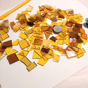 May include: A collection of amber and yellow stained glass pieces in various shapes and sizes. A yellow pencil, a silver coin, and a glass cutter are also present. The glass pieces are scattered on a white surface, suggesting a craft project.
