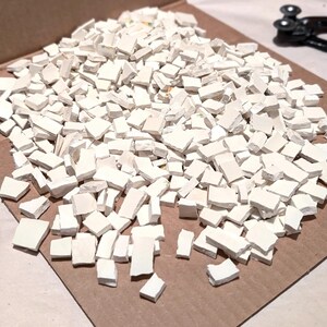 May include: A large collection of small, irregular, matte white ceramic mosaic tiles scattered across a surface. A pair of blue-handled tile nippers and a silver-colored coin are positioned near the pile of white ceramic pieces for scale.
