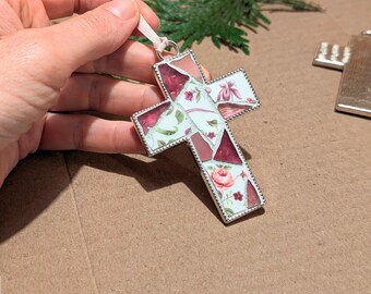 May include: A white and pink floral mosaic cross ornament with a silver border. The cross is hanging from a white ribbon.
