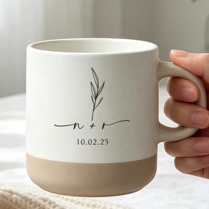 May include: A two-toned ceramic mug with a speckled white upper section and a solid tan base. The mug features a black line drawing of a sprig, the lowercase letters n + r, and the date 10.02.25 printed in a simple, dark font on the front.