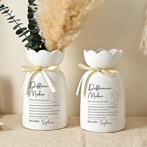 Può includere: Due vasi in ceramica bianca con bordi smerlati e nastri color crema. I vasi hanno il testo "Difference Maker" e sono riempiti con erba della pampa secca e verde. Una tazza di caffè è su un piattino.