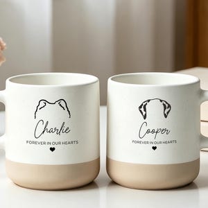 Puede incluir: Dos tazas de cerámica blanca con acabado moteado y base color canela. Cada taza presenta una ilustración de líneas negras de orejas de perro, un nombre en cursiva y el texto "FOREVER IN OUR HEARTS" junto a un pequeño icono de corazón.