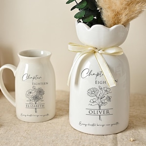 May include: A white ceramic mug and vase with floral designs and text. The mug reads "Chapter Eighteen" and "Elizabeth". The vase reads "Chapter Eighteen" and "Oliver". Both have a floral bouquet illustration and a ribbon tied around the vase.