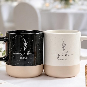 May include: Two ceramic mugs with a speckled finish and a solid base. The black mug features the text m + b and 11.21.25 with a leaf graphic. The white mug features the text g + h and 09.07.25 with a leaf graphic. Both have a handle on the side.