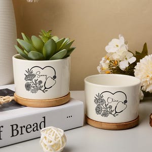 May include: Two cream ceramic plant pots with bamboo saucers. One pot contains a succulent. Each pot has a black floral design with a stethoscope and a name. The text "Sunny" and "Olivia" are visible.