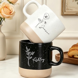 May include: Two ceramic mugs with speckled finishes and tan bases. The top white mug features a black floral line drawing and the name Jeanne. The bottom black mug features a white floral line drawing and the name Ashley, both with handles.