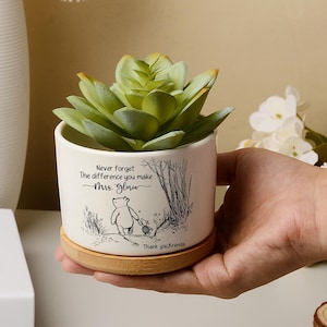 May include: A white ceramic plant pot with a faux succulent, featuring a Winnie the Pooh illustration and the text "Never forget the difference you make, Mrs. Glow". The pot sits on a wooden base, ideal for home decor.
