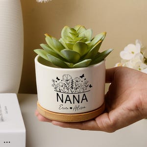 Peut inclure: Pot de fleurs en céramique blanche avec une plante succulente artificielle. Le pot porte l'inscription "NANA" en noir, avec "Eric & Alison" en dessous. Il repose sur une base ronde en bois, pour une petite plante.