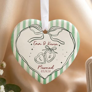 May include: A heart-shaped ceramic ornament with a white ribbon, featuring light green vertical stripes and a central illustration of two small hearts with the initials E and K, the text Emm & Kevin, Married, and the date 03.26.26 in dark red.