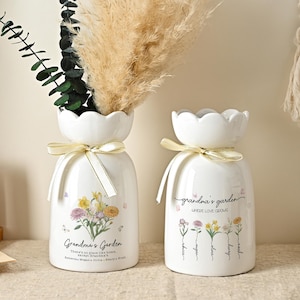 May include: Two white ceramic vases with scalloped rims and cream-coloured ribbon bows. The vases feature floral illustrations and the text "Grandma's Garden" with "WHERE LOVE GROWS" printed on the surface, containing dried pampas grass and eucalyptus.