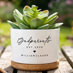 May include: A white ceramic planter with a wooden base containing a green succulent. The planter features black text reading Godparents EST. 2026 with a small heart and the name WILLIAM CLAUDE. Small gardening tools and a burlap bag are nearby.