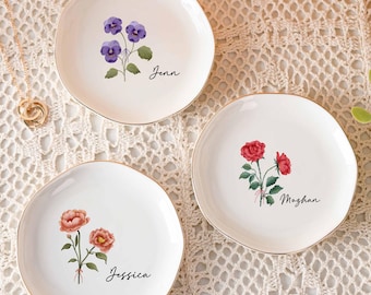 Könnte beinhalten: Drei runde weiße Keramikschalen mit goldfarbenen Rändern. Jede Schale zeigt eine individuelle Blumenillustration und einen personalisierten Namen in schwarzer Schreibschrift: violette Stiefmütterchen für Jenn, rote Rosen für Meghan und orangefarbene Blumen für Jessica.