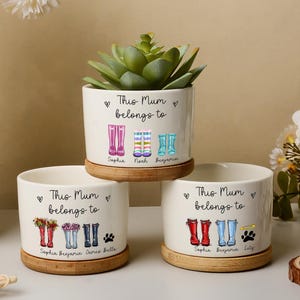 Peut inclure: Trois pots de fleurs en céramique blanche avec des soucoupes en bambou, chacun portant l'inscription "This Mum belongs to" et des illustrations colorées de bottes de pluie. Un pot contient une plante succulente. Des noms sont imprimés sous les bottes.