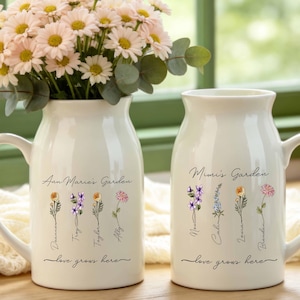 May include: Two white ceramic pitchers with handles, each featuring a floral graphic and the text "love grows here." One pitcher is labelled "Ann Marie's Garden" with four flower stems, and the other is "Mimi's Garden" with four flower stems.