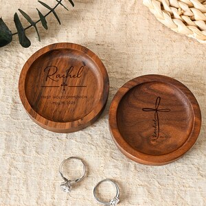 Puede incluir: Dos platos redondos de madera para anillos con texto grabado y un diseño de cruz. Uno de los platos dice "Rachel, Primera Comunión, 18 de mayo de 2025". También hay dos anillos de plata con piedras transparentes.