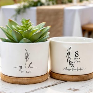 Op de afbeelding: Twee cilindrische plantenbakken van wit keramiek met houten onderzetters. Eén bevat een groene vetplant en is bedrukt met g + h en 09.11.25. De andere is leeg en toont een bladillustratie met de tekst 8 years of us en Maya & Hudson.
