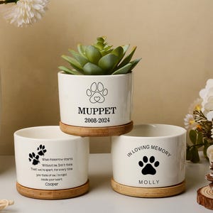 Peut inclure: Trois pots de fleurs en céramique blanche avec des soucoupes en bambou. L'un des pots a une plante succulente et un motif de pattes avec le nom "Muppet" et des dates. Un autre a une empreinte de patte et le nom "Cooper". Le troisième a une empreinte de patte et le nom "Molly".