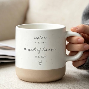 May include: A white ceramic mug with a speckled finish and a tan base. The text reads "sister EST. 1993", "maid of honor", "EST. 2026", and a small hand-drawn heart. The mug features a curved handle and a smooth, cylindrical body design.