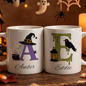 Puede incluir: Dos tazas de cerámica blanca con diseños de Halloween. La taza izquierda muestra una letra A morada con un sombrero de bruja, telaraña y caldero, etiquetada como Amber. La derecha muestra una letra E verde con una linterna y un cuervo, como Eddie.