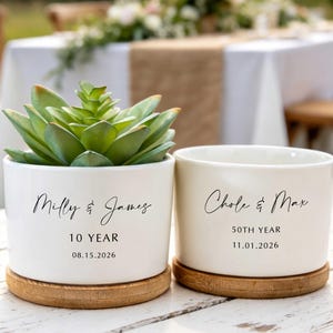 Peut inclure: Deux pots de fleurs cylindriques en céramique blanche avec des bases en bois. Le pot de gauche contient une plante succulente verte et porte le texte Milly & James, 10 YEAR, 08.15.2026. Le pot de droite porte le texte Chole & Max, 50TH YEAR, 11.01.2026.