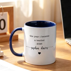 Op de afbeelding: Een witte keramische mok met een donkerblauwe binnenkant en handvat. De buitenkant bevat zwarte tekst: the year i became a teacher 2026, gevolgd door de naam Sophia Davis in een sierlijk lettertype en een klein zwart hartje eronder.