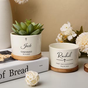 Puede incluir: Dos macetas de cerámica blanquecina con posavasos de madera, una con una suculenta y la otra vacía. La primera maceta tiene el nombre "Jack", una cruz y la fecha "03.14.2025". La segunda maceta tiene el nombre "Rachel", una cruz y la fecha "May 18, 2025".