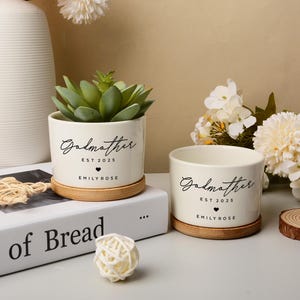 May include: Two cream ceramic plant pots with the word "Godmother" in script, a heart, and "Emily Rose" printed in black. One pot contains a succulent. Both sit on wooden coasters. The text "EST 2025" is also printed on the pots.