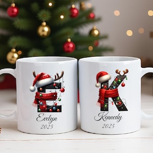 Pode incluir: Duas canecas de cerâmica branca com designs natalinos. Uma caneca apresenta a letra "E" com um gorro de Papai Noel, chifres de rena e um cachecol vermelho. A outra caneca tem a letra "K" com decorações semelhantes. Ambas as canecas têm nomes e o ano de 2025.