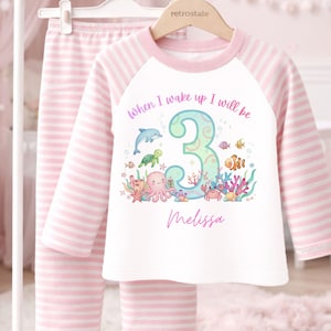 May include: A pink and white striped pajama set. The white long-sleeved top has a colourful underwater graphic with the number 3, the text "When I wake up I will be", and a name. The set is hanging on a white rack.