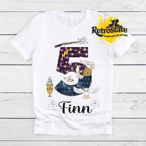 Könnte beinhalten: Ein weißes Kurzarm-T-Shirt mit einer großen dunkelblauen Zahl fünf, die mit goldenen Sternen und einem Blitz verziert ist. Das Design enthält einen Zauberstab, einen goldenen geflügelten Schlüssel, einen Kelch und eine weiße Eule. Der Name Finn steht darunter.