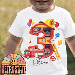 May include: A white t-shirt featuring a large red number three shaped like a fire station. The design includes a yellow firefighter helmet, a red fire engine, a dalmatian, a ladder, a fire hose, and colourful balloons. The name Oliver is printed below.