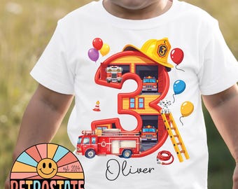 May include: A white t-shirt featuring a large red number three shaped like a fire station. The design includes a yellow firefighter helmet, a red fire truck, a dalmatian, a ladder, a fire hose, and colorful balloons. The name Oliver is printed below.