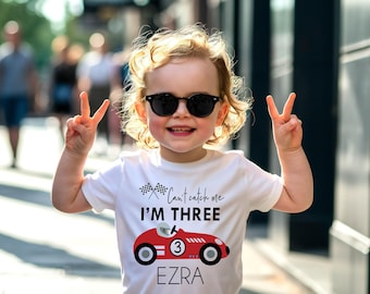 May include: A white short-sleeved t-shirt featuring a graphic of a red race car with the number three, two checkered flags, and the text "Can't catch me I'M THREE" above the car and the name "EZRA" printed in black lettering below the car.