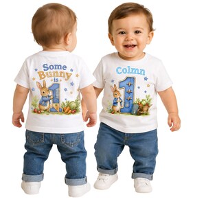 May include: Two white short-sleeved t-shirts featuring a Peter Rabbit graphic, a large blue number one, and garden vegetables. One shirt displays the text Some Bunny is 1, while the other shirt displays the name Colmn above the number one.