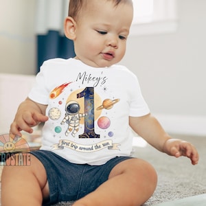 May include: A white short-sleeve t-shirt featuring a space-themed graphic with an astronaut, planets, and a large number one. The text reads Mikey's and first trip around the sun. The design includes a comet, stars, and a yellow sun background.