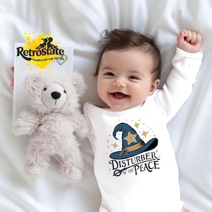 May include: A white long-sleeved baby bodysuit featuring a graphic of a blue wizard hat with a gold star and the text "DISTURBER OF THE PEACE" in black lettering, accompanied by a small, fluffy, light grey teddy bear toy.