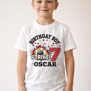 May include: A white t-shirt with a bowling-themed graphic. The shirt features the text "BIRTHDAY BOY STRIKE! OSCAR" with a bowling pin and ball design, and the number 7 in red. The boy is wearing blue jeans.