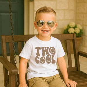 May include: A white short-sleeved t-shirt featuring the brown text "TWO COOL" in a wavy, retro-inspired font. The letters "O" in the text are replaced with smiling face icons. The shirt is worn with tan shorts and silver-framed aviator sunglasses.
