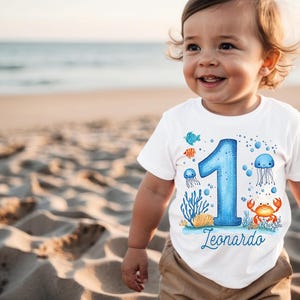 May include: A white short-sleeved t-shirt featuring a large blue number one graphic surrounded by colourful illustrations of a crab, jellyfish, and coral. The name Leonardo is printed in blue script below the number on the front of the shirt.