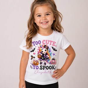 May include: A white short-sleeved t-shirt featuring a graphic of a rainbow-maned unicorn dressed in a black outfit, surrounded by Halloween elements like bats, pumpkins, and ghosts. The text reads "TOO CUTE TO SPOOK" with the name "Elizabeth" below.