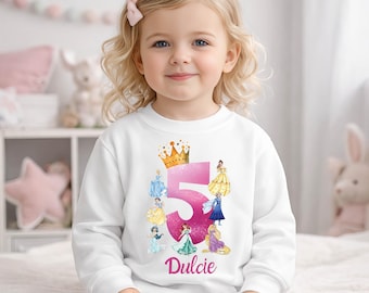 Puede incluir: Sudadera blanca con un gran número 5 rosa, una corona dorada e ilustraciones de princesas. El nombre "Dulcie" está impreso debajo. La sudadera es para un niño.