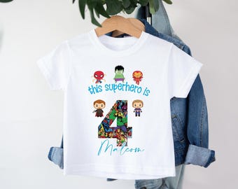 May include: White t-shirt featuring the text "this superhero is 4 Malcolm" in blue and teal script. The number 4 is filled with comic book images. Superhero cartoon characters are above the text.
