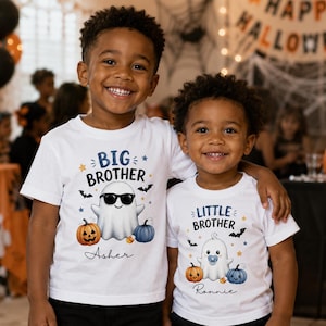 Könnte beinhalten: Zwei weiße kurzärmelige T-Shirts mit Halloween-Motiven. Das linke Shirt zeigt einen Geist mit Sonnenbrille, Kürbisse und den Namen Asher sowie den Schriftzug BIG BROTHER. Das rechte Shirt zeigt einen Geist mit Schnuller, Kürbisse und den Namen Ronnie sowie den Schriftzug LITTLE BROTHER.