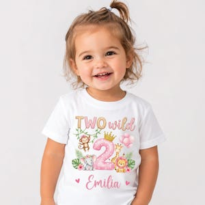 May include: A white short-sleeved t-shirt featuring a graphic print with the text TWO wild and the name Emilia. The design includes a large pink polka-dot number two, a gold crown, pink balloons, and illustrations of a monkey, elephant, giraffe, and lion.