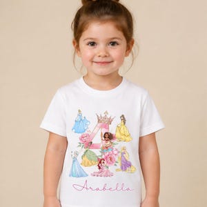 May include: A white short-sleeved t-shirt featuring a large pink watercolour number 4 topped with a gold crown. The design includes various illustrated princesses, pink floral accents, and the name Arabella written in a cursive pink script font.