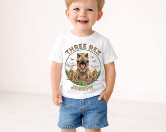 May include: A white short-sleeved t-shirt featuring a graphic of a Tyrannosaurus Rex inside a circular frame with volcanoes, tropical plants, and the text "THREE REX" at the top and the name "JOSEPH" at the bottom in brown lettering.