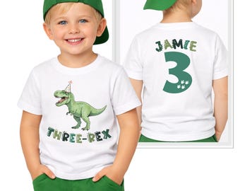 May include: A white short-sleeved t-shirt featuring a green T-Rex wearing a party hat with the text "THREE-REX" below it. The back of the shirt displays the name "JAMIE" above a large green number "3" with small dinosaur footprint graphics.