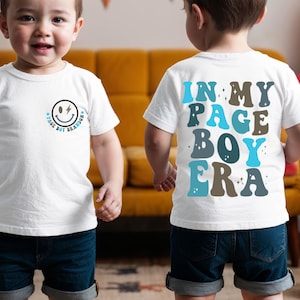 May include: Two young children wearing white t-shirts and denim shorts. One shirt features a smiley face with lightning bolts and the text "PAGE BOY BRANDON". The other shirt says "IN MY PAGE BOY ERA" in blue and brown letters.